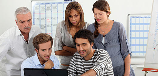 fünf Personen schauen gemeinsam in einen Laptop. Eine Person zeigt auf den Bildschirm
